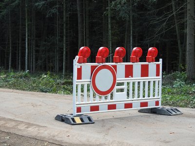 Das Bild zeigt eine Straßensperre, die mit einem Verkehrszeichen „Durchfahrt verboten“ (ein roter Kreis mit einem weißen Innenfeld) versehen ist. Die Sperre besteht aus einer Barriere in Rot und Weiß, die auf einer Straße oder einem Weg aufgestellt ist. Oben auf der Barriere befinden sich mehrere rot leuchtende Warnlampen, die die Sperre bei Dunkelheit oder schlechten Sichtverhältnissen deutlicher machen sollen.