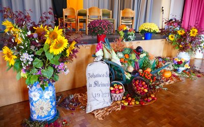 Auf dem Bild sieht man die Bühne des Bürgersaales im Bürgerhauses. Die Bühne und der Bereich davor sind herbstlich und farbenfroh geschmückt mit Blumen, Früchten, Gemüsen, die sich unter anderem auf einem blauen Schubkarren befinden. Am linken Bildrand steht eine große Bodenvase gefüllt mit Sonnenblumen, daneben ein weißer Getreidesack mit grauer Aufschrift.